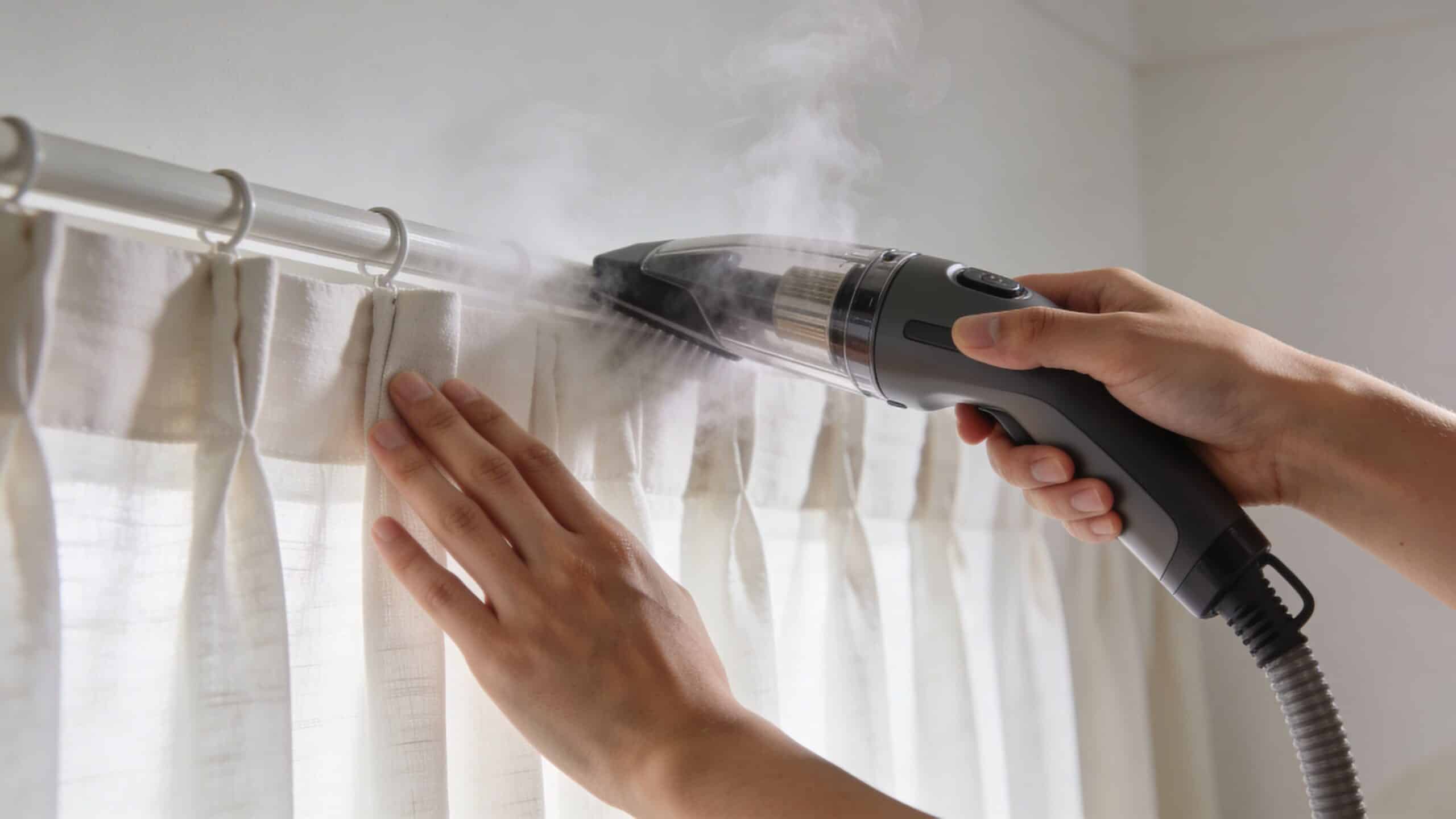 A person uses a handheld steam cleaner to refresh and sanitize white fabric curtains at home.