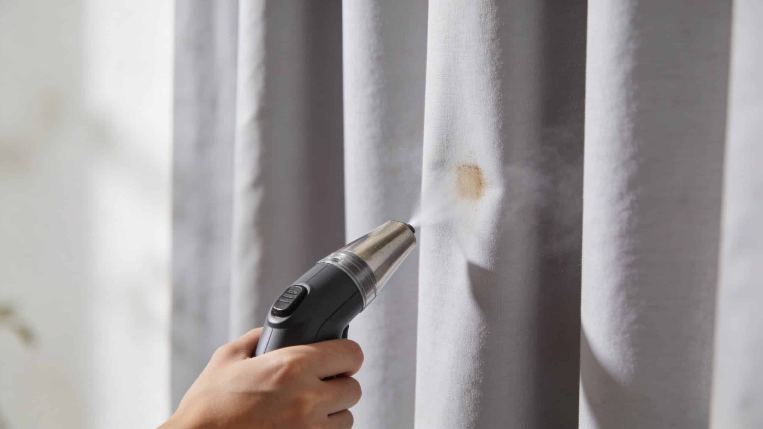 A person uses a handheld portable steam cleaner to remove a stain from light gray curtains indoors.
