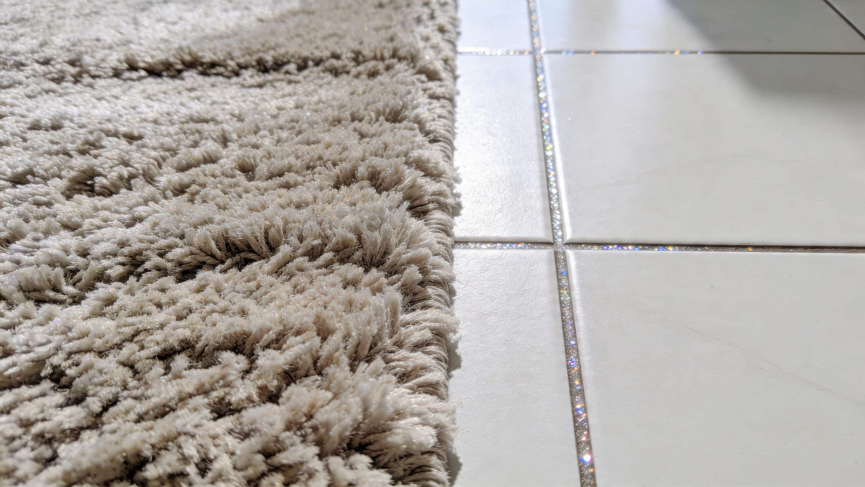 A close-up view of a shaggy beige carpet edge resting next to clean white floor tiles.