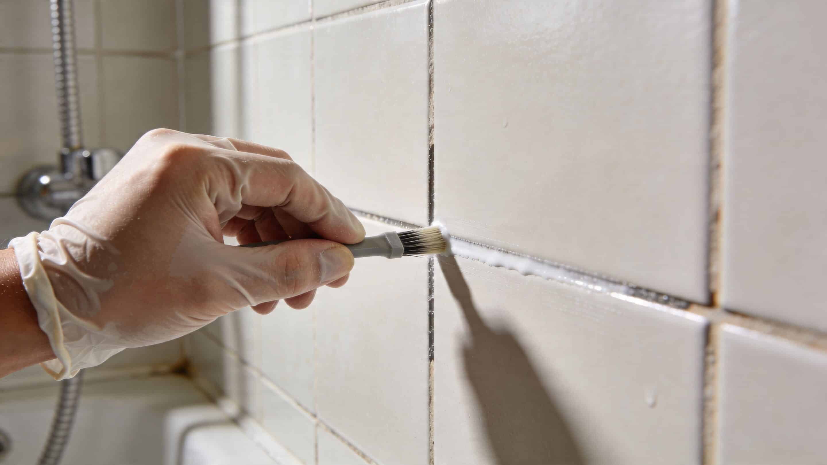 A hand wearing a protective glove using a brush to apply sealant to bathroom tile grout.