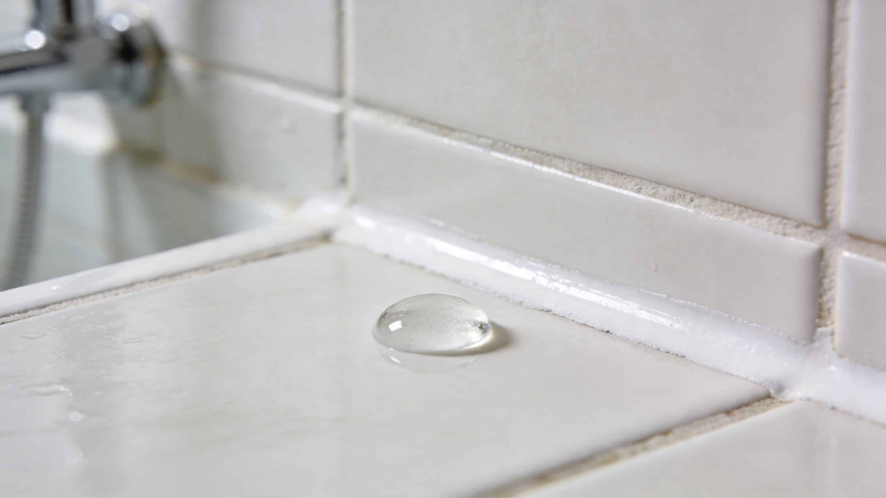 A single crystal clear water droplet sitting on a clean, white tiled bathroom surface with grout.