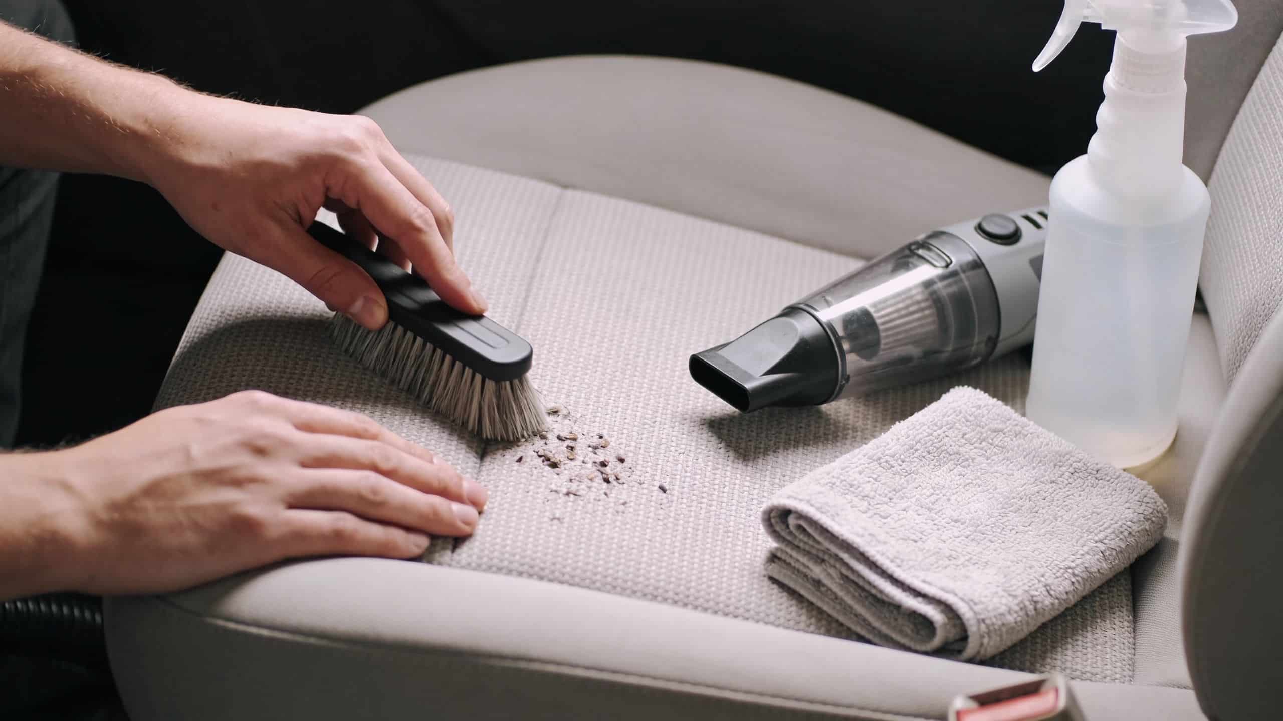 A person using a brush to clean loose dirt and debris from a light-colored car seat.