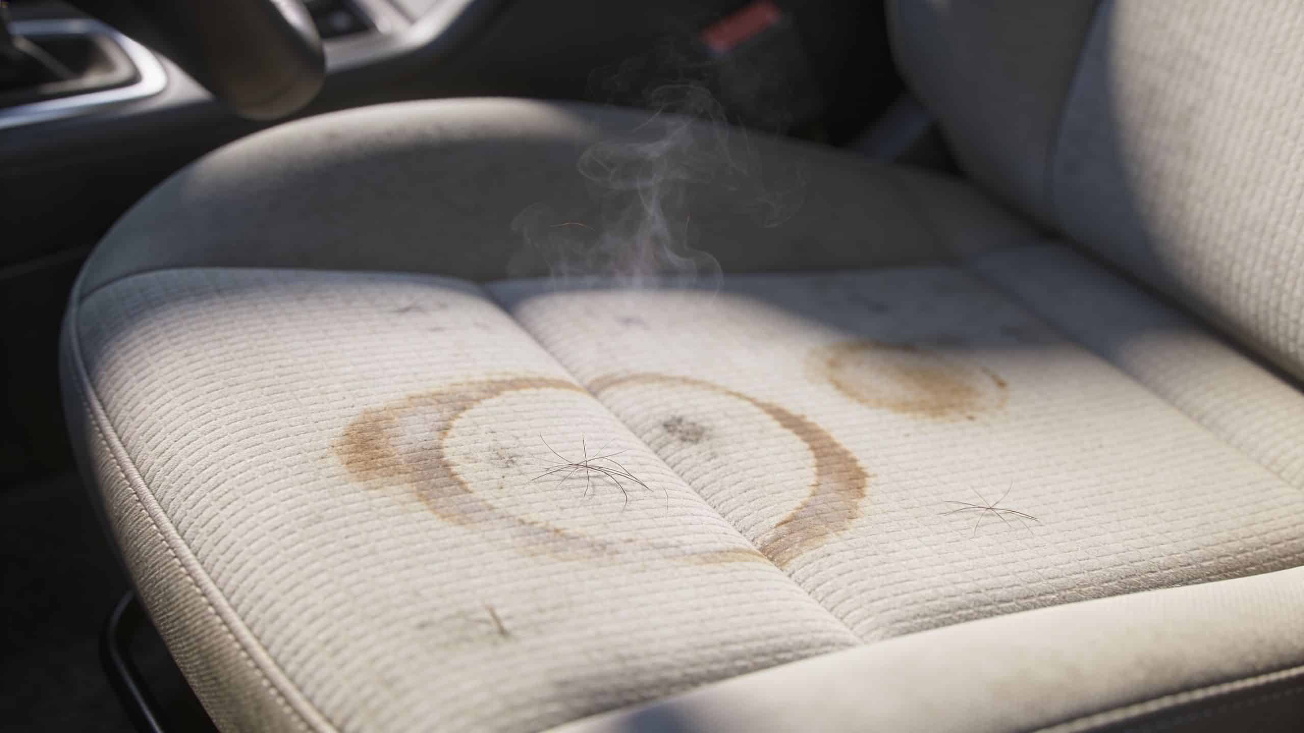 A car seat made of fabric with visible brown circular coffee stains and steam rising from it.