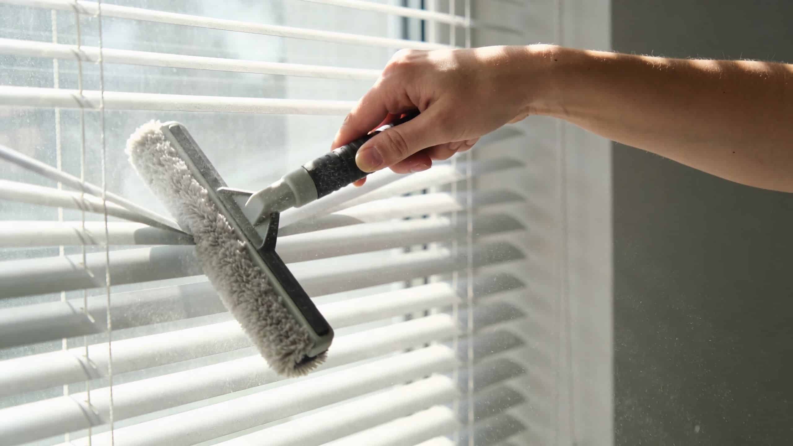 A hand using a specialized microfiber tool to dust and clean white venetian blinds on a window.